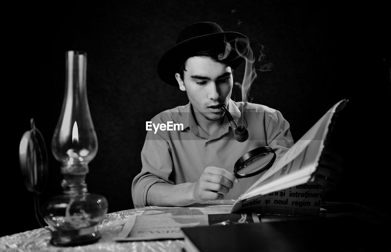 Young man smoking pipe while using magnifying glass at table