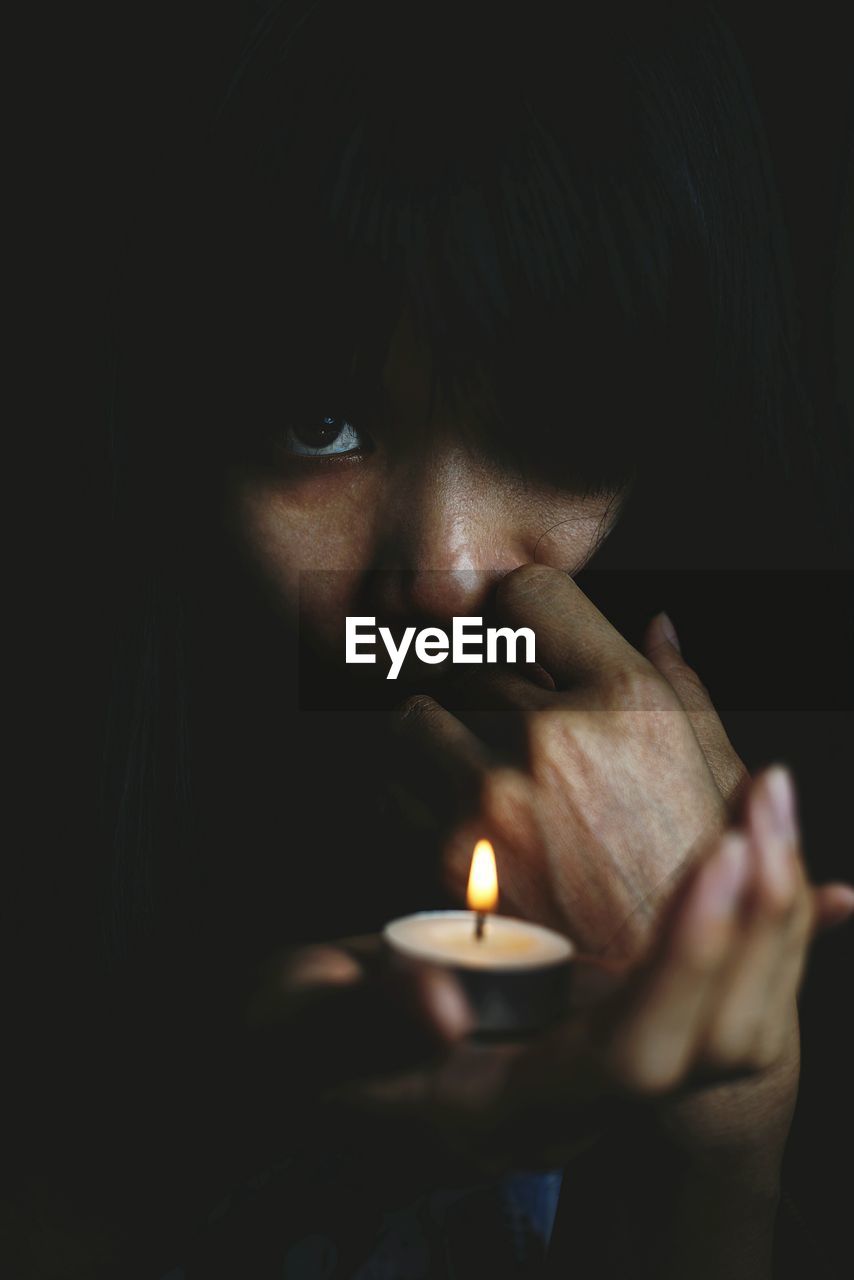 Close-up portrait of woman holding lit tea light against black background