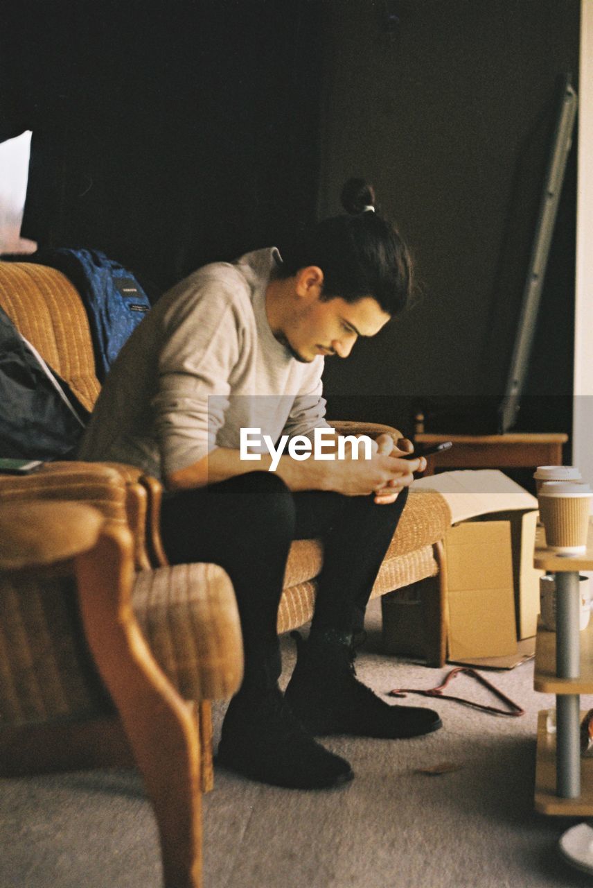 Side view of young man using phone while sitting on sofa at home