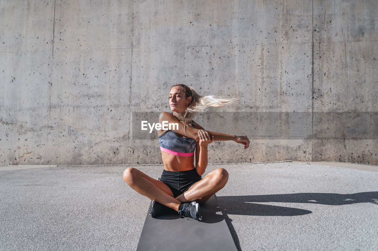 Slim female athlete with eyes closed in sportswear sitting on mat and stretching arms during workout in city