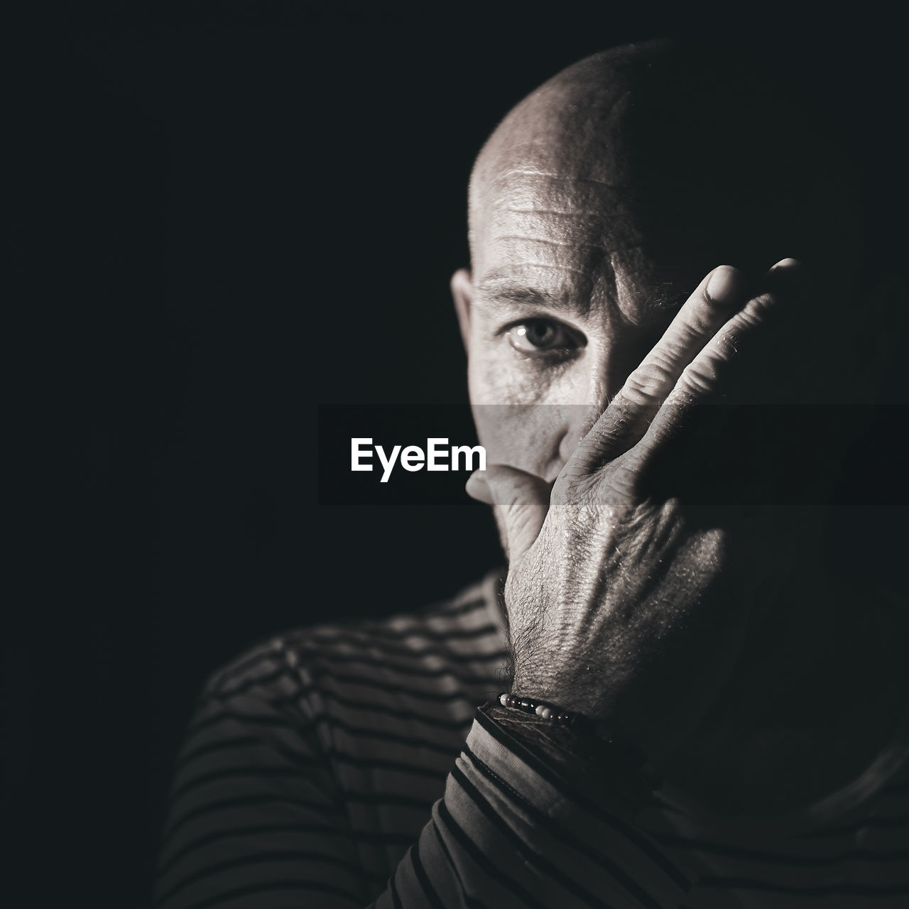 Close-up portrait of bald man covering mouth against black background