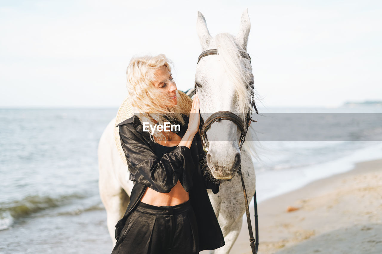 Young blonde woman in black clothes riding white horse on seascape background