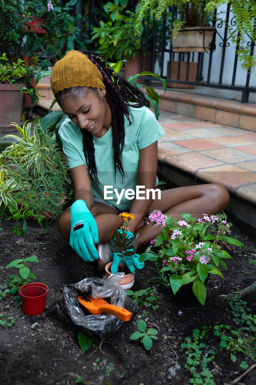 From above of smiling black female gardener sitting on ground in hothouse and transplanting kalanchoe flower