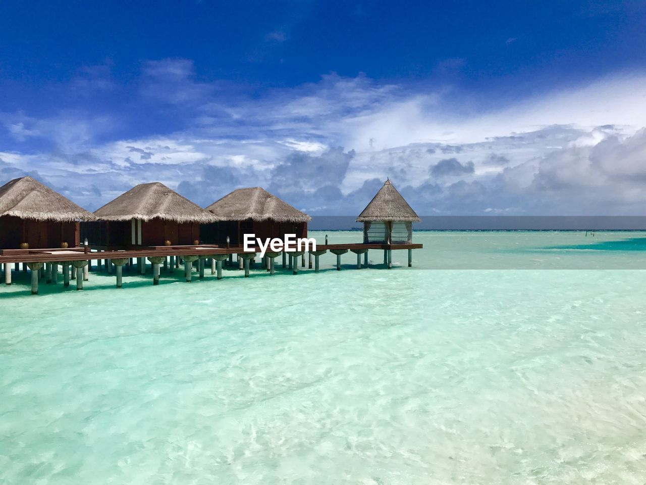 water, sky, hut, ocean, sea, thatched roof, nature, land, architecture, scenics - nature, beauty in nature, built structure, stilt house, cloud, house, travel destinations, beach, blue, vacation, tranquility, building, building exterior, tranquil scene, roof, turquoise colored, shore, no people, tourist resort, lagoon, idyllic, tropical climate, holiday, landscape, trip, travel, island, residential district, resort, outdoors, environment, day, coast, beach hut, sand, bay, tourism, mountain, sunlight, summer, horizon, holiday villa