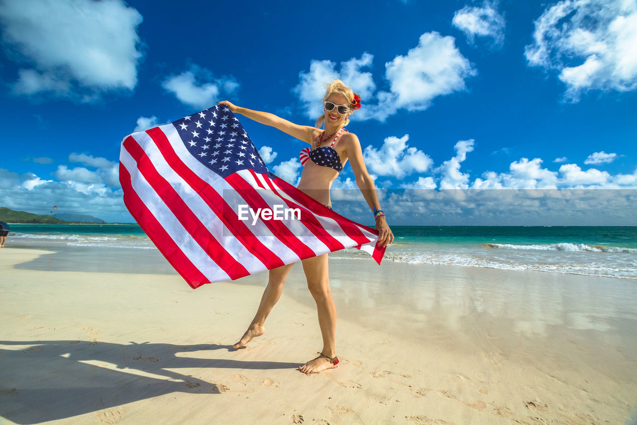 Portrait of happy woman holding american flag while standing at beach against blue sky