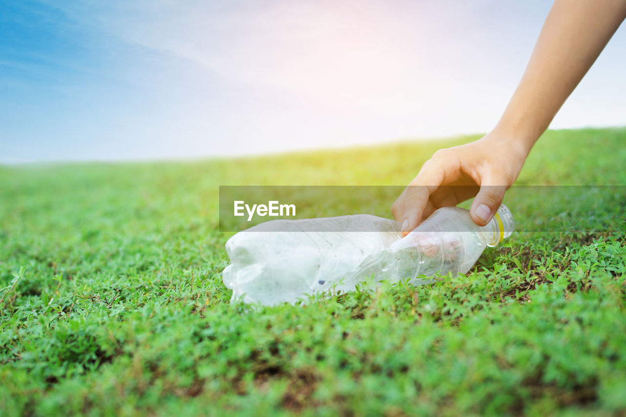 Cropped hand holding plastic bottle on grassy field against sky