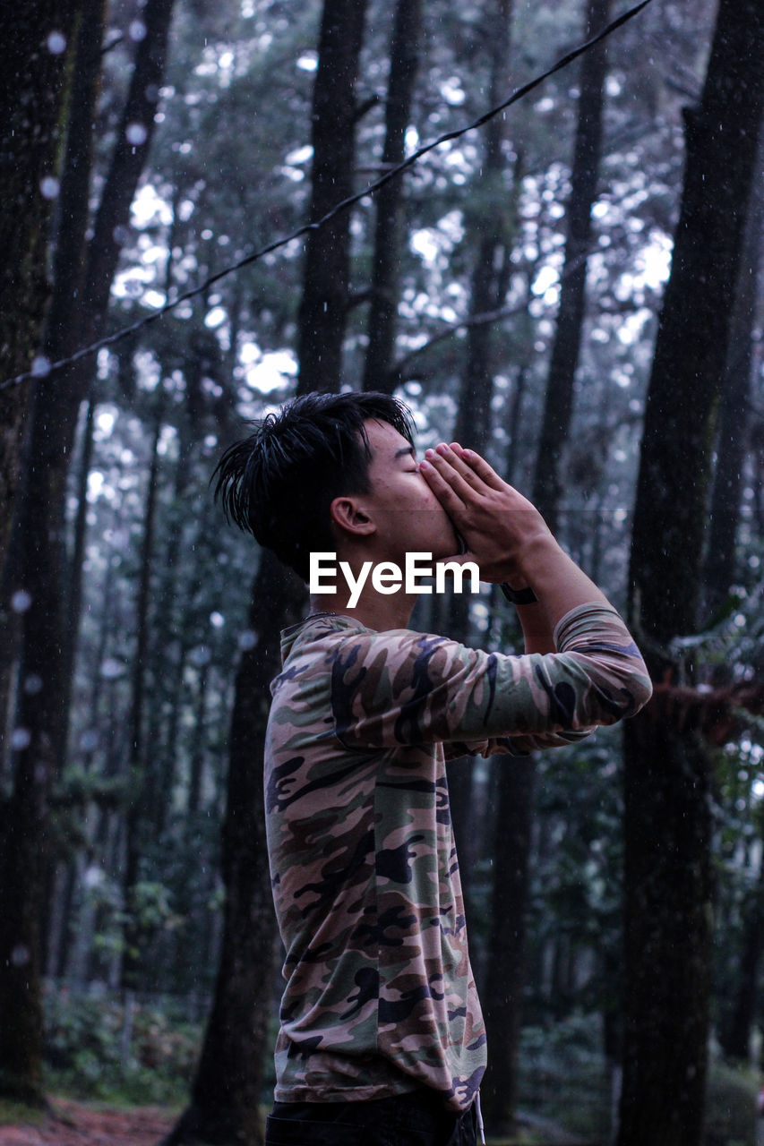 Side view of young man standing against trees in forest