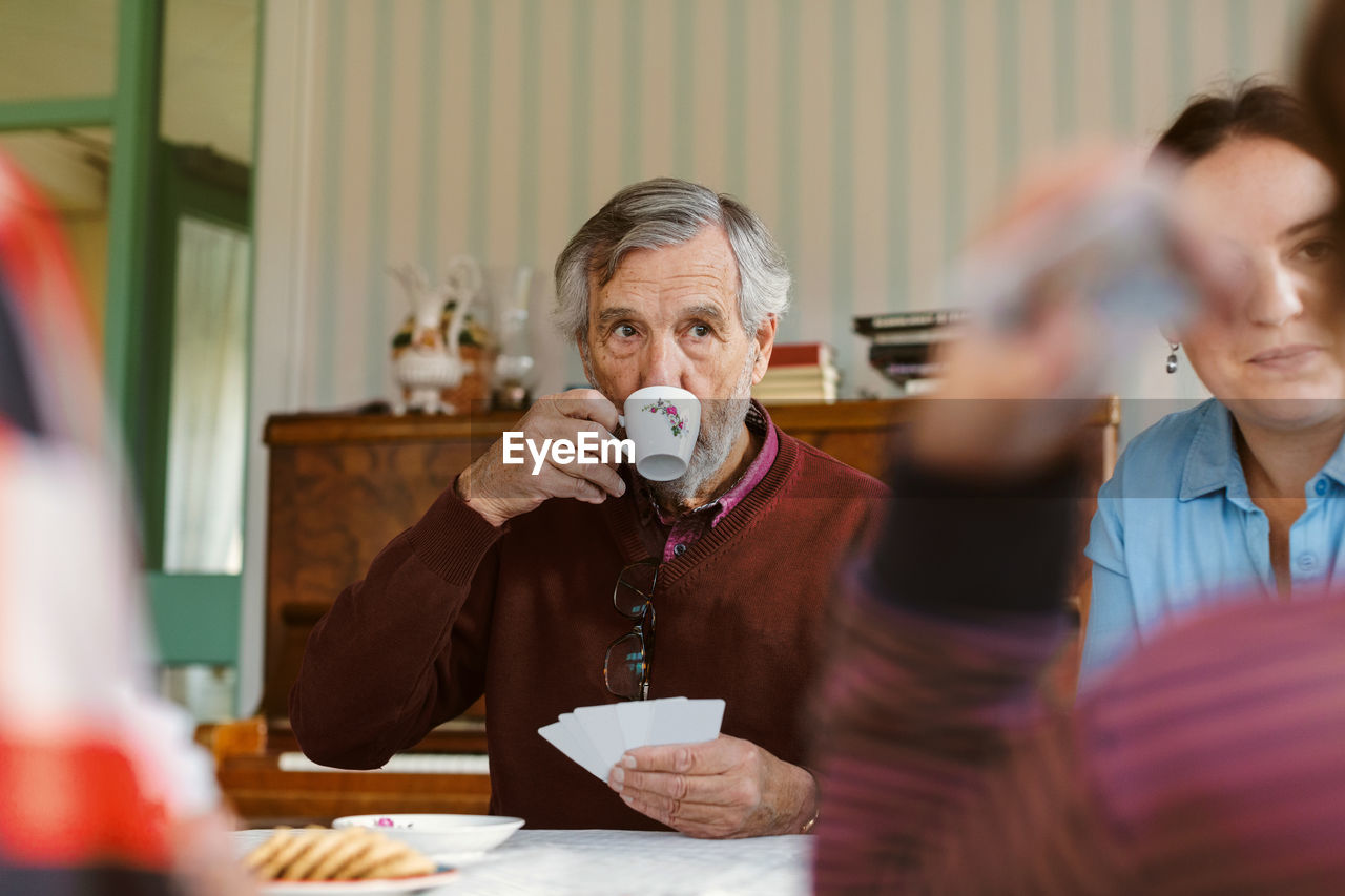 Senior man drinking coffee while playing cards with family at home