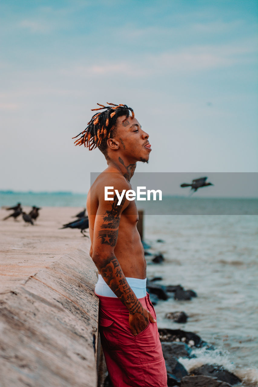 Side view of shirtless man standing at beach against sky