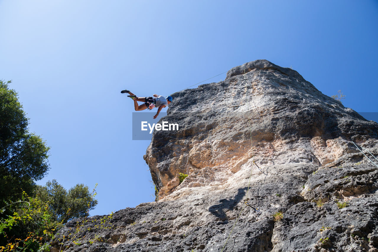 Low angle view of man rock climbing against clear blue sky