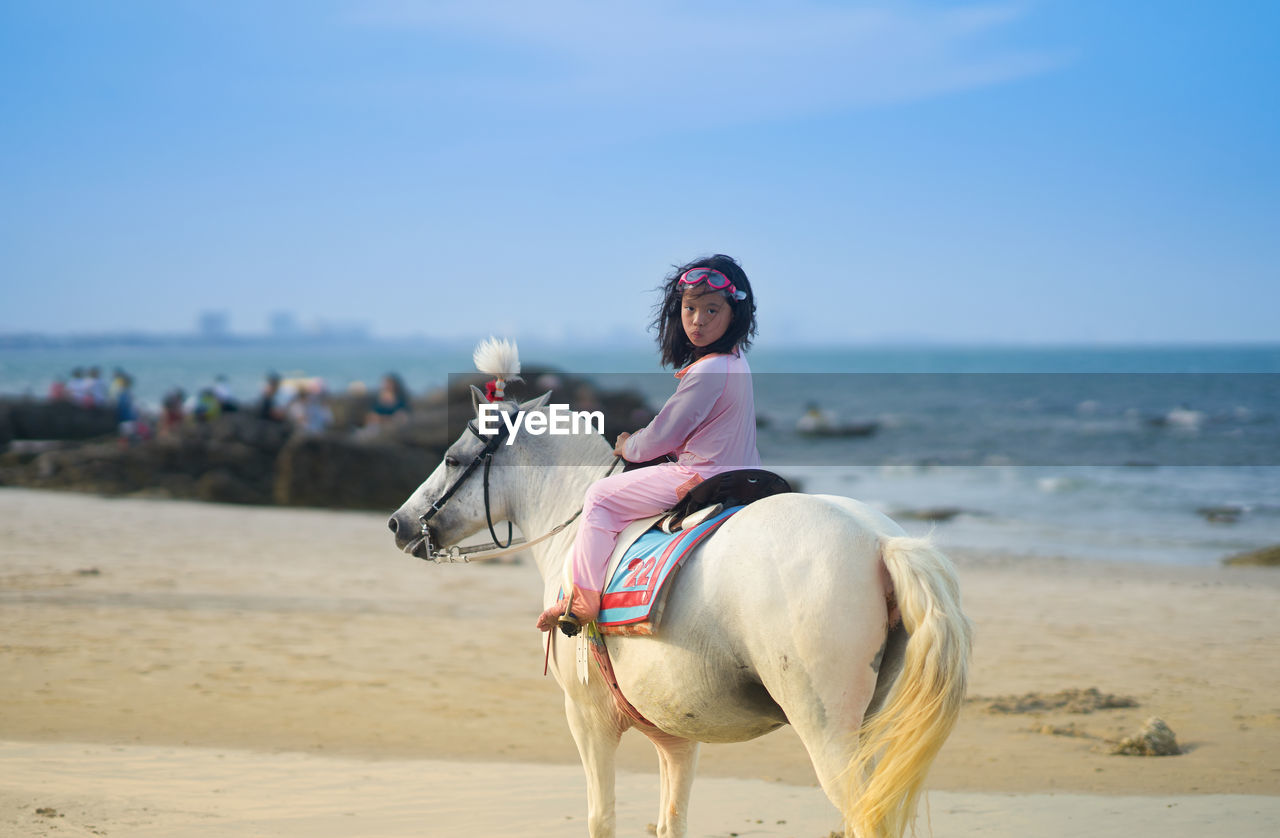 Full length of girl sitting on horse at beach against sky