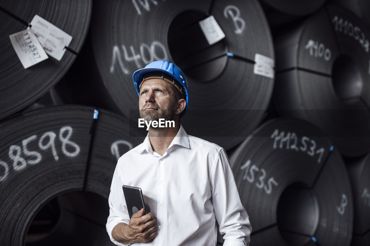 Male entrepreneur holding digital tablet looking up while standing against steel rolls in factory