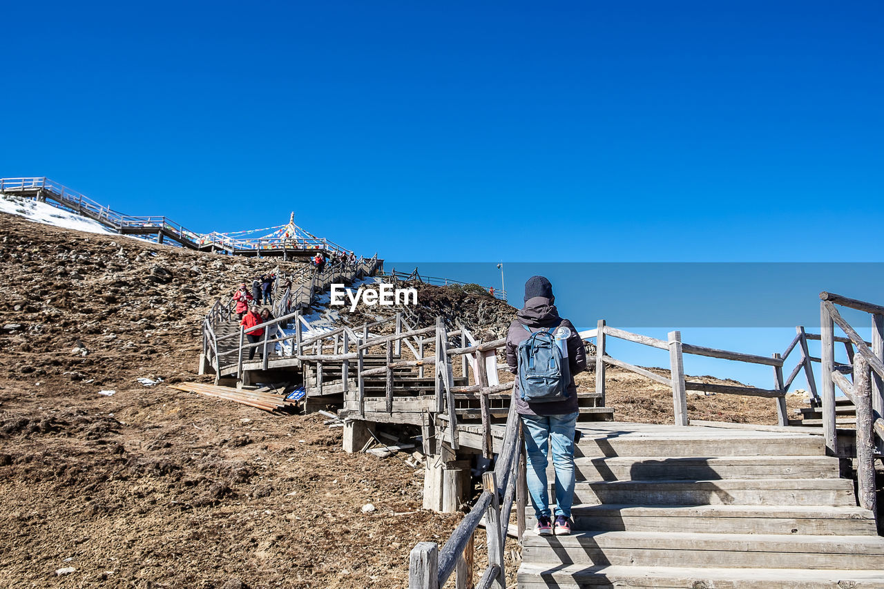 REAR VIEW OF PEOPLE STANDING ON STAIRCASE AGAINST BLUE SKY