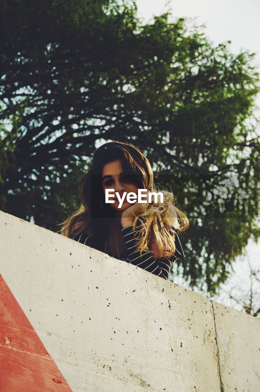 Low angle portrait of smiling girl with hand on chin standing by retaining wall against tree