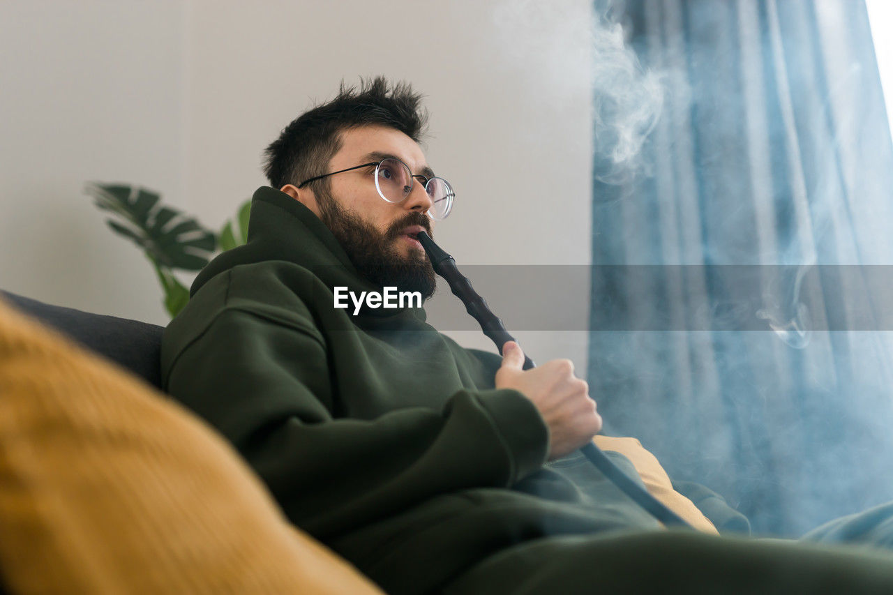adult, conversation, men, eyeglasses, one person, glasses, person, sitting, indoors, beard, facial hair, communication, activity, looking, furniture, relaxation, smoke, clothing, human face, portrait, contemplation, lifestyles, young adult, copy space, black hair, casual clothing, holding, business, selective focus, serious, technology