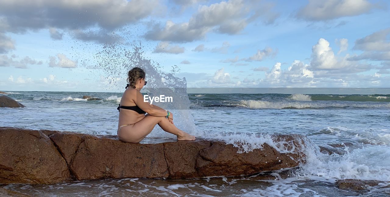 Side view of woman sitting on rock in sea against sky