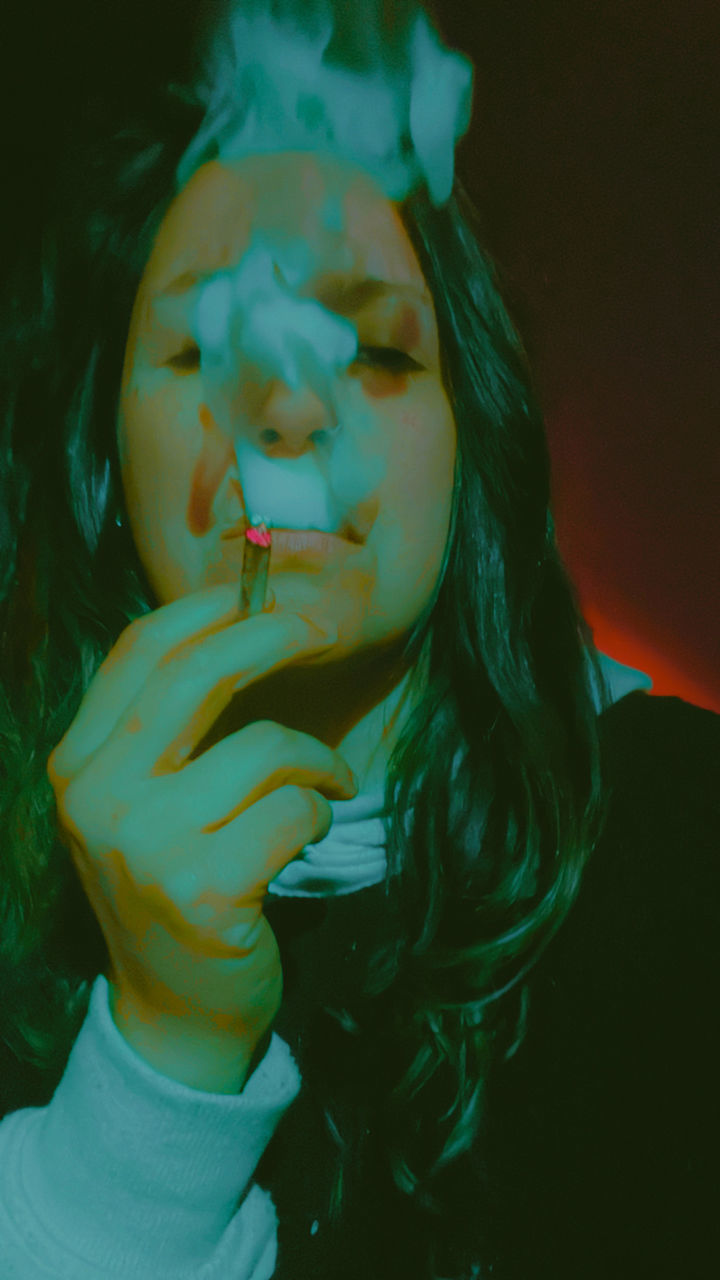 one person, blue, portrait, green, women, young adult, headshot, adult, front view, indoors, lifestyles, bad habit, human face, looking at camera, leisure activity, smoking issues, close-up, person, eyes closed, long hair, hairstyle, darkness, cigarette, female, smoking, smoke