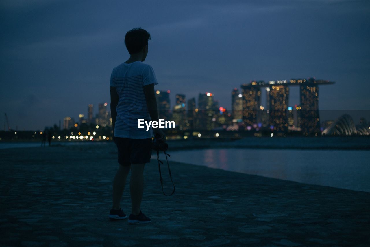 Full length rear view of man standing by illuminated marina bay sands at dusk