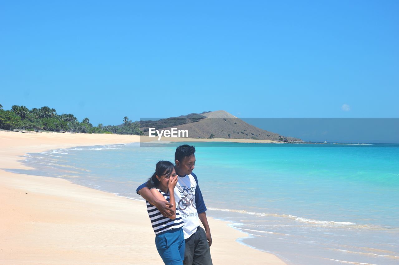 Young couple standing at beach against clear blue sky