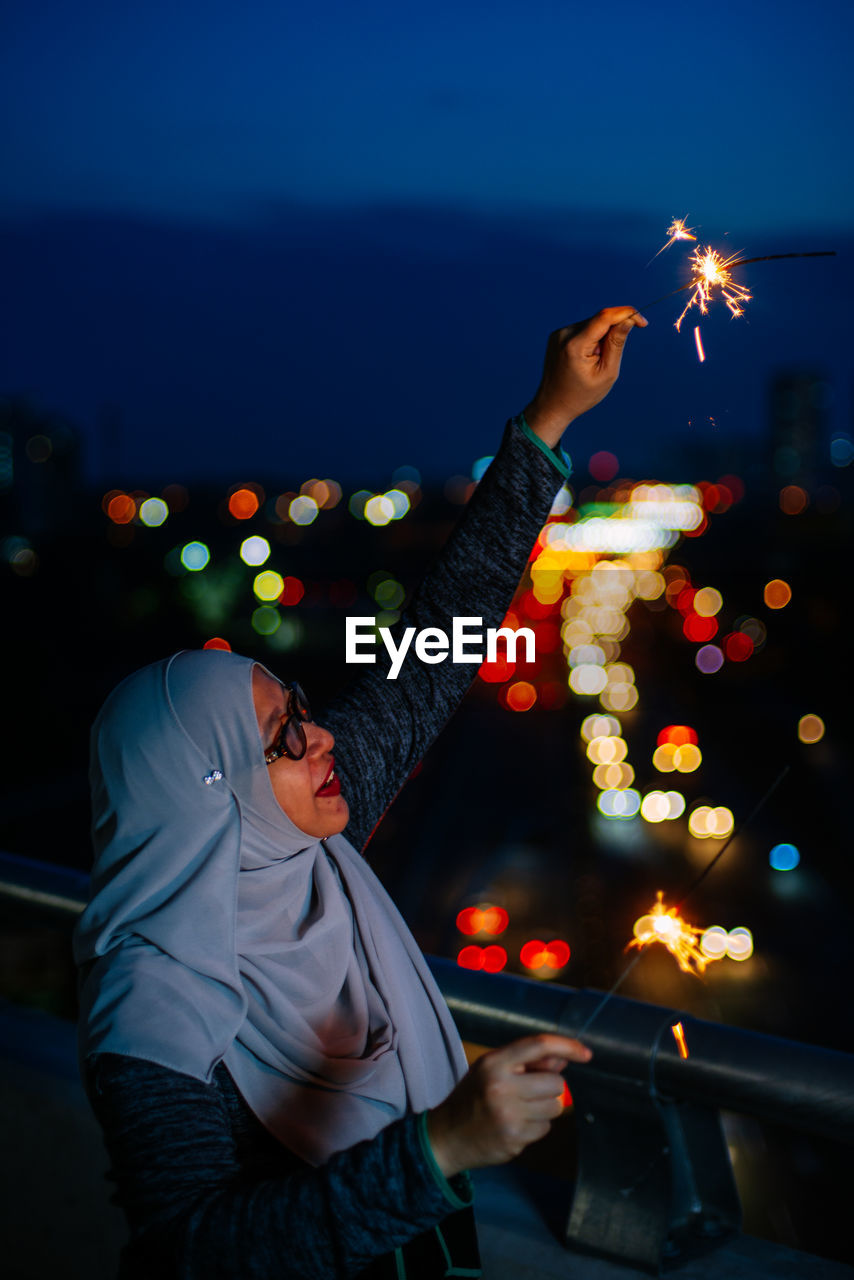 Side view of smiling mid adult woman holding burning sparkler at night