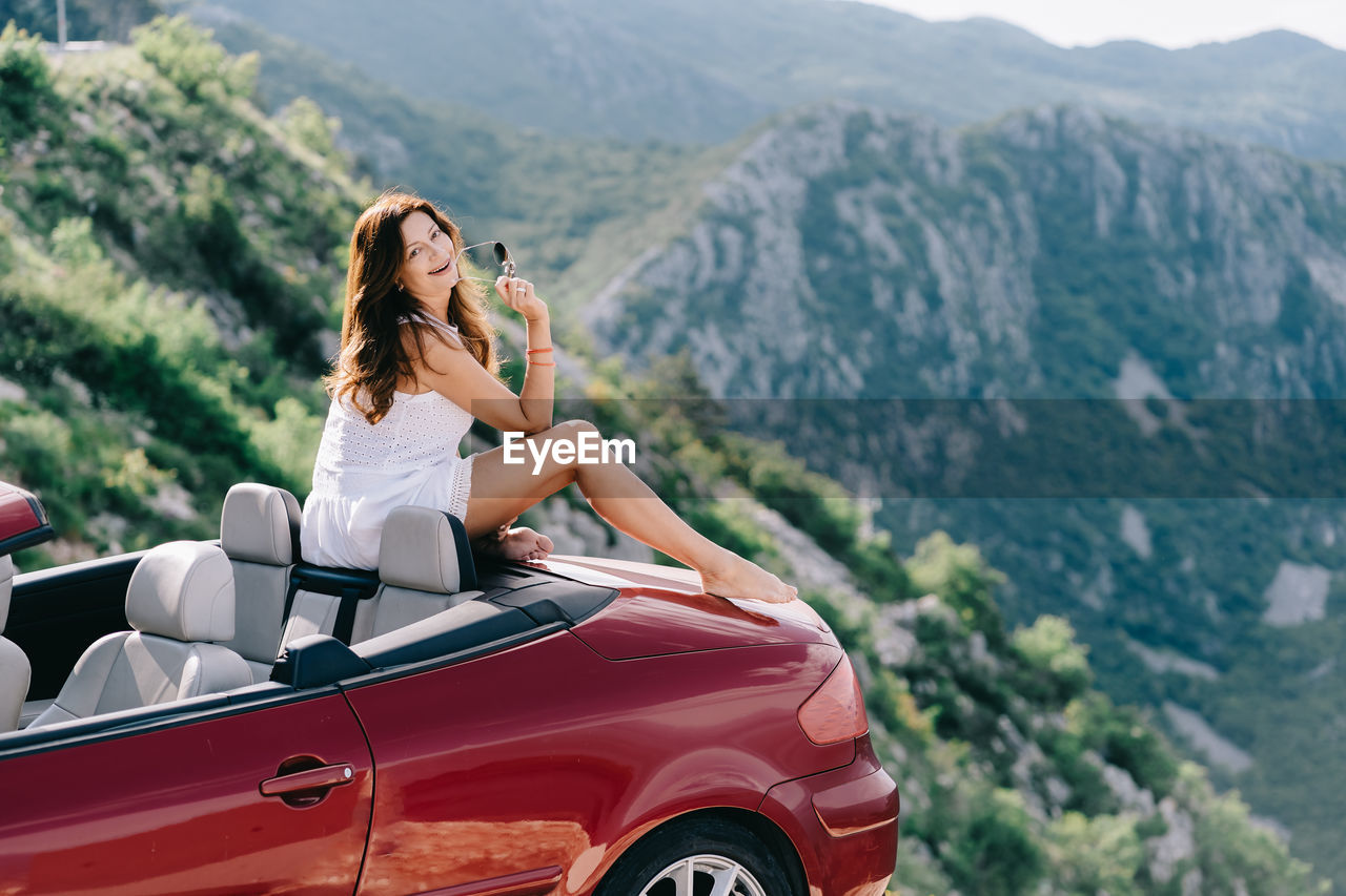 REAR VIEW OF WOMAN SITTING ON CAR AGAINST MOUNTAIN
