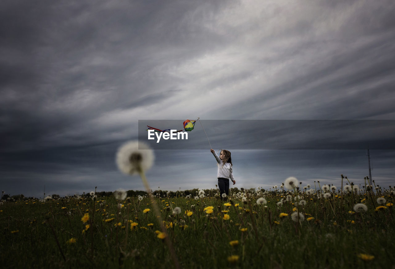Girl playing with kite while standing on dandelion field against stormy clouds