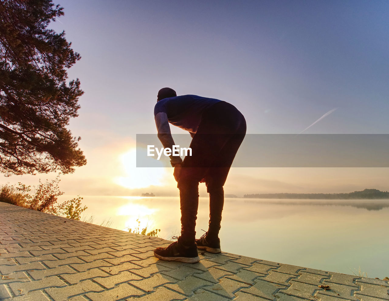 Short break for breath. silhouette of active man exercising and stretching on lake beach at sunrise