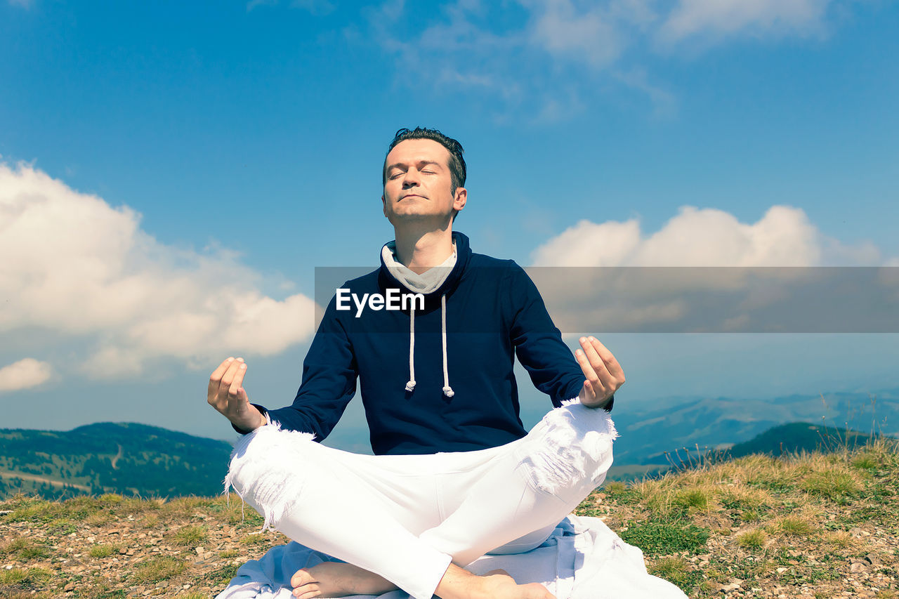 Mid adult man in lotus pose practicing yoga on a field at mountain peak. copy space.