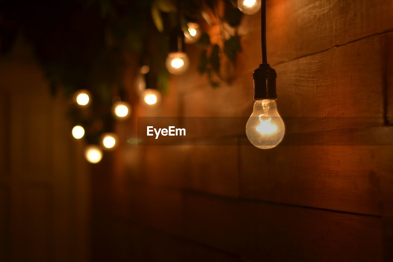 Close-up of illuminated light bulbs hanging from ceiling by wooden wall