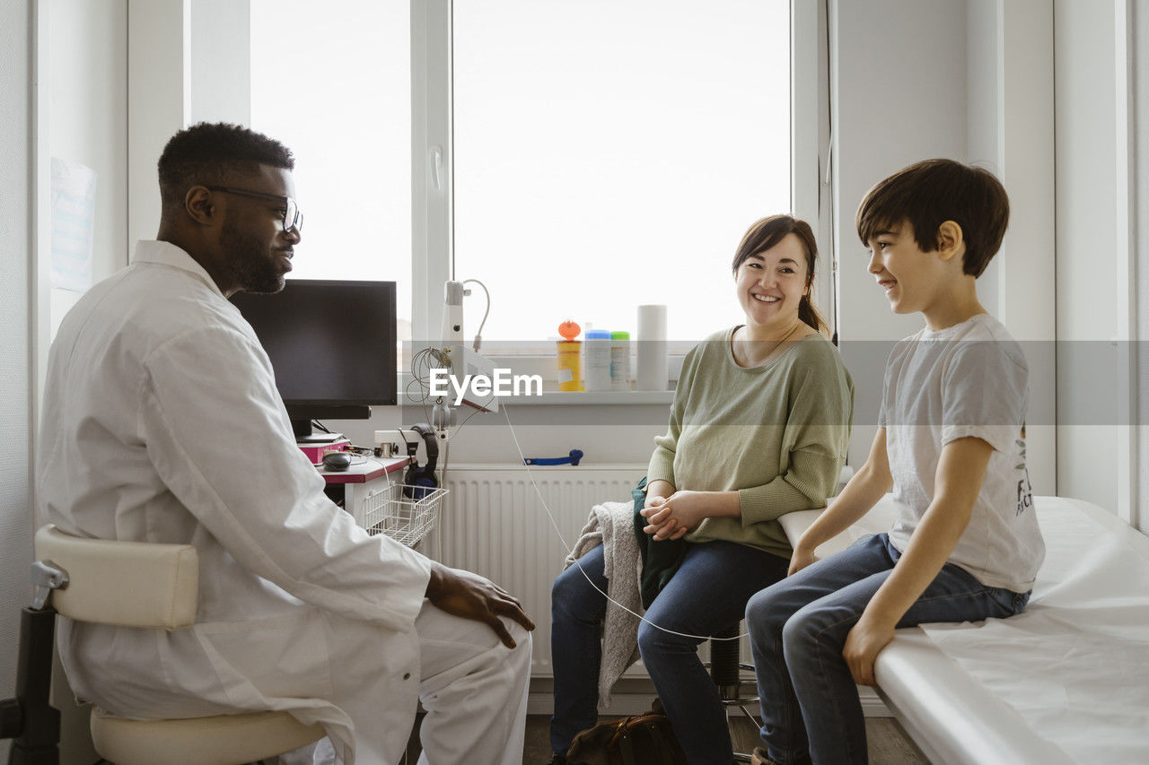 Smiling boy by mother discussing with male pediatrician in examination room at healthcare center