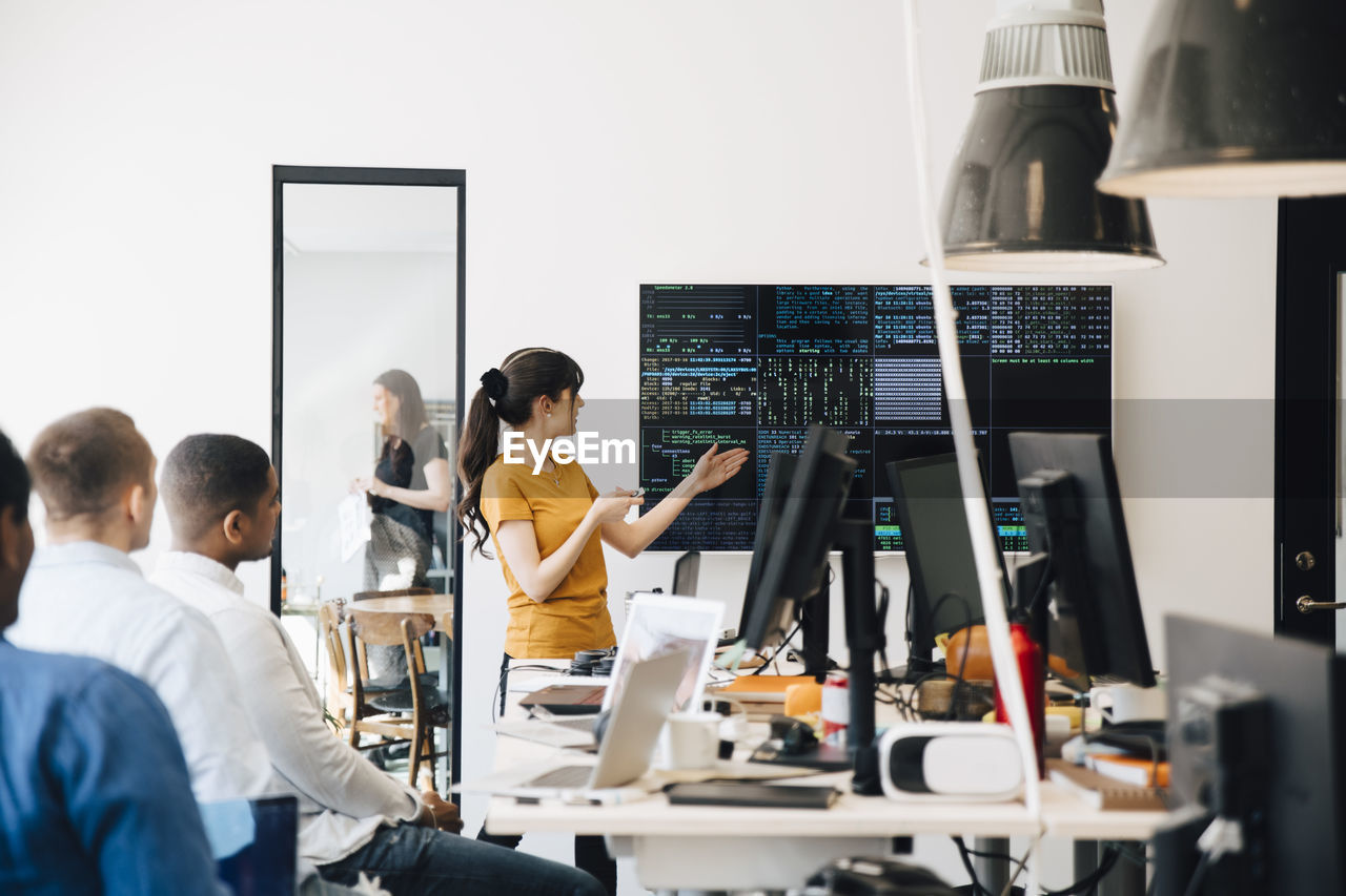 Female computer hacker presenting code on television screen during meeting with programmers in creative office