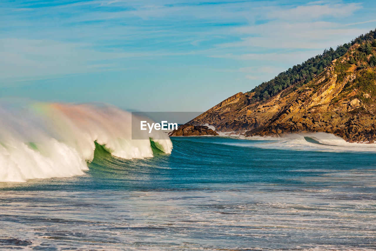 Large rainbow wave in sea against sky with rocky headland in background.