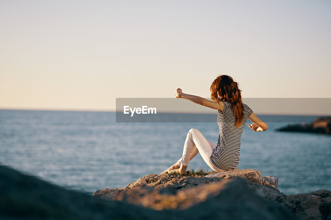 WOMAN BY ROCK ON SEA AGAINST CLEAR SKY