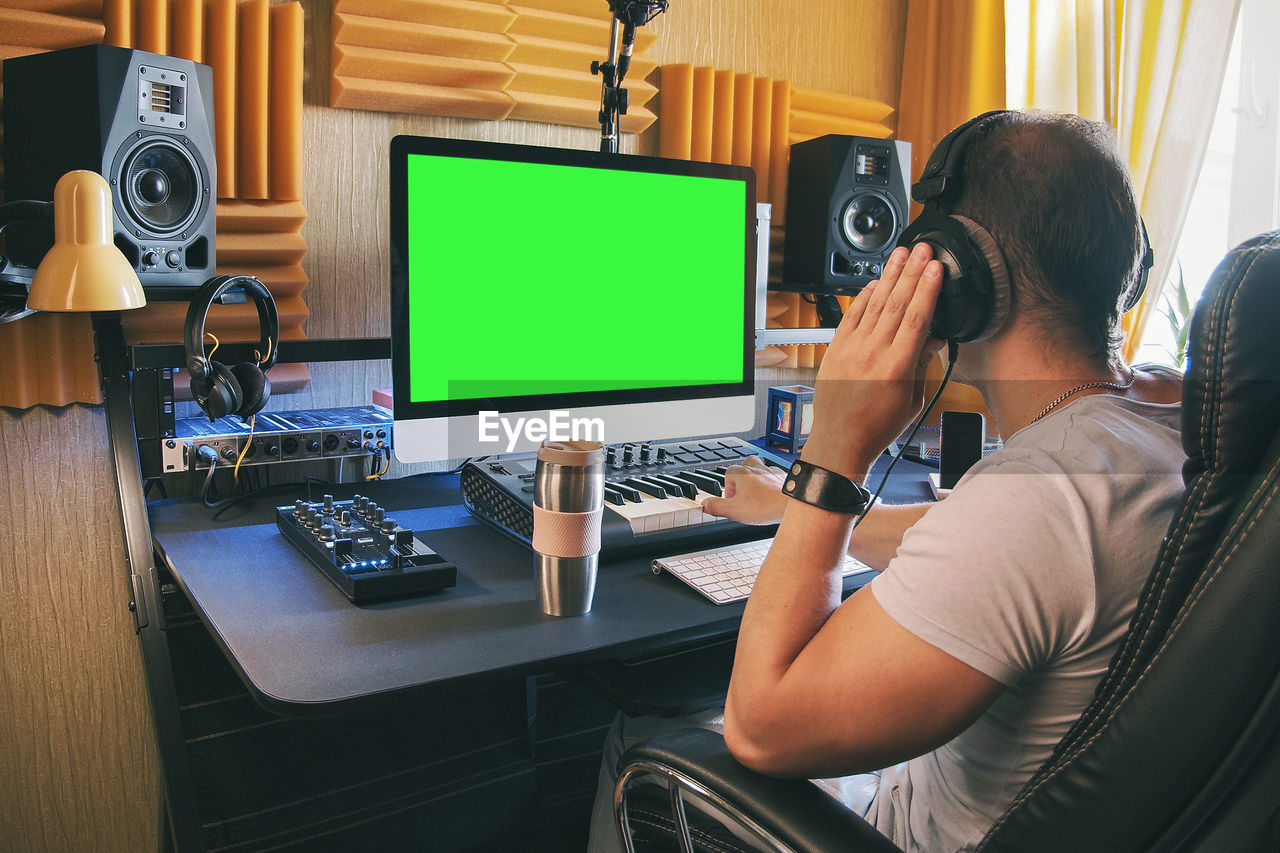 A man produce electronic music in home studio using headphones and look in green screen on monitor