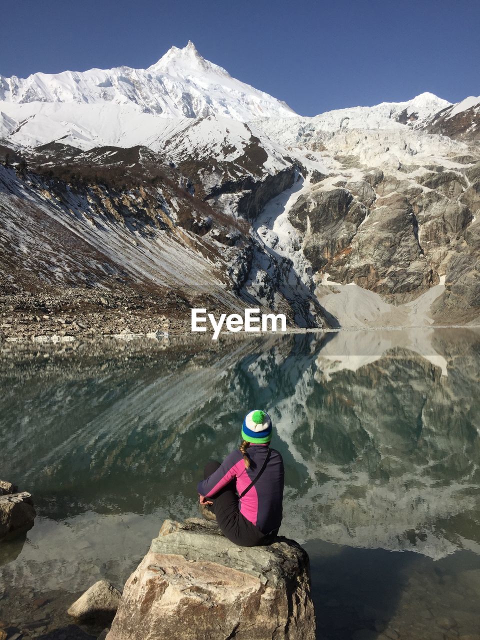 Rear view of woman sitting on rock by lake with snowcapped mountains reflection