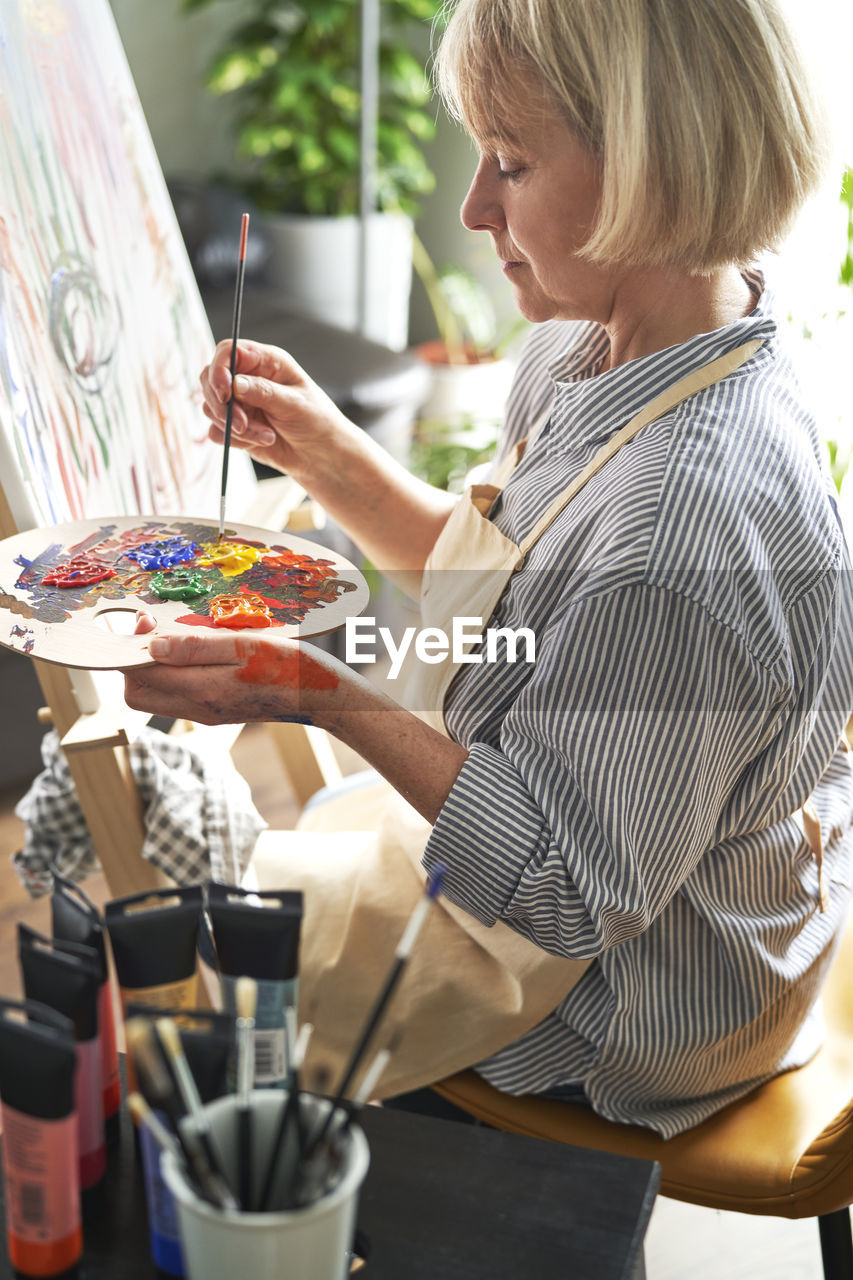 Blond woman with paintbrush mixing colors on palette in front of easel at home