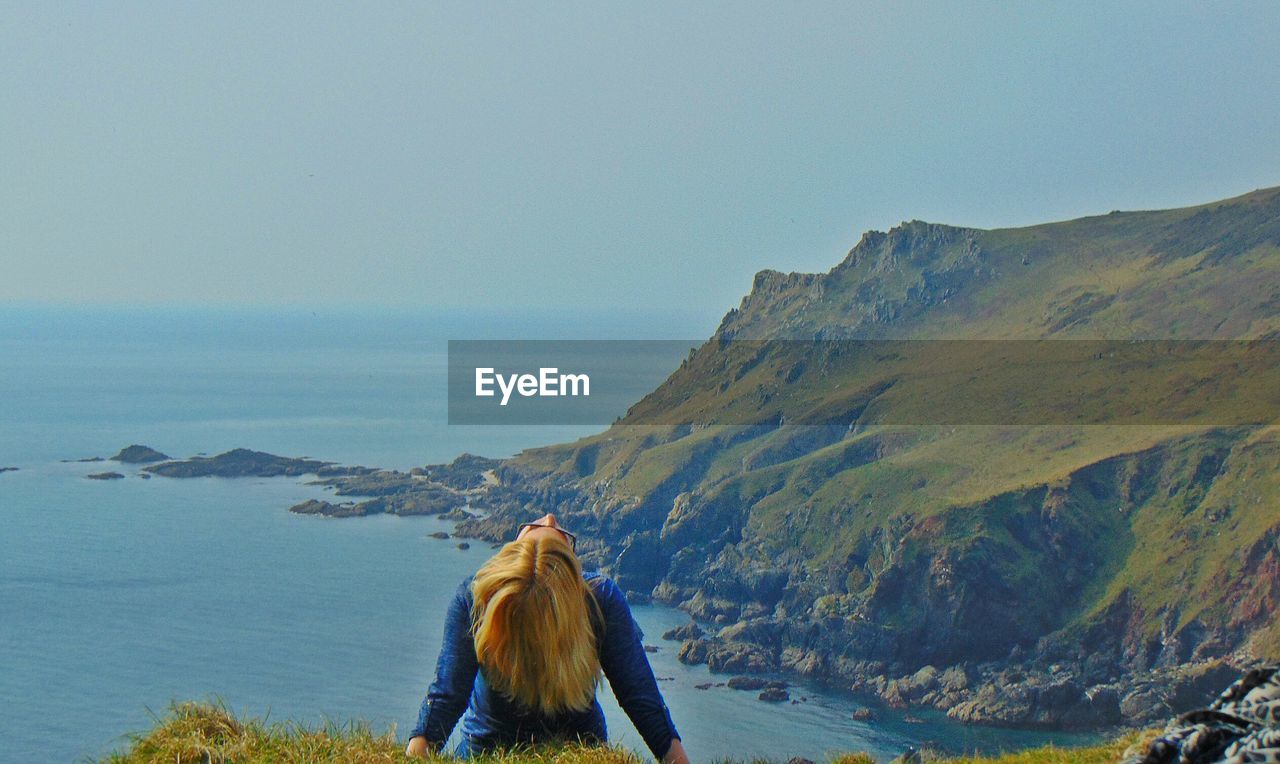 Rear view of woman tossing hair on cliff by sea against clear sky