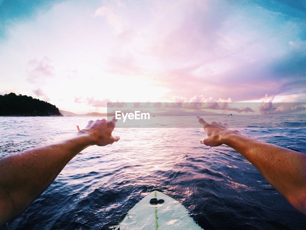 Cropped hands of man over surfboard in sea against sky during sunset