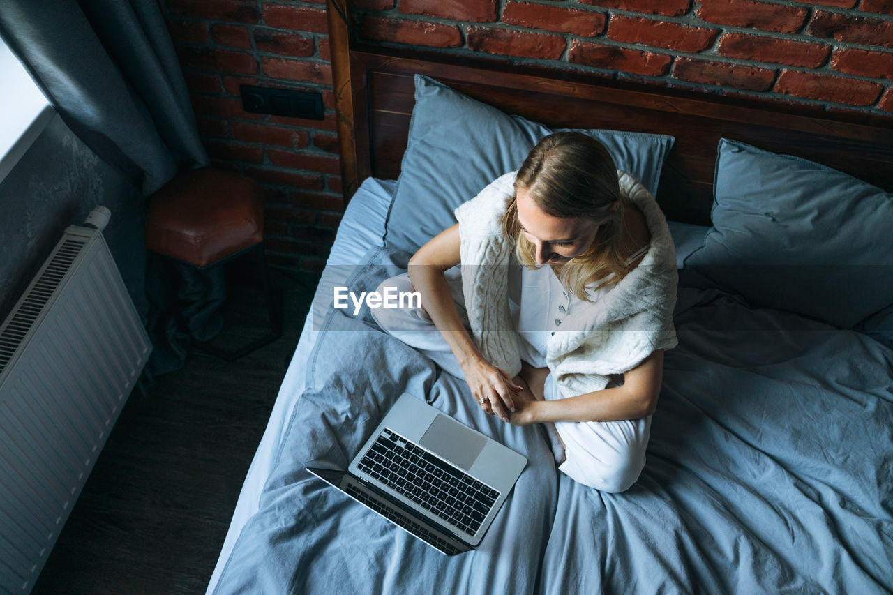 Adult woman freelancer with blonde long hair in casual clothes using laptop sitting on bed 