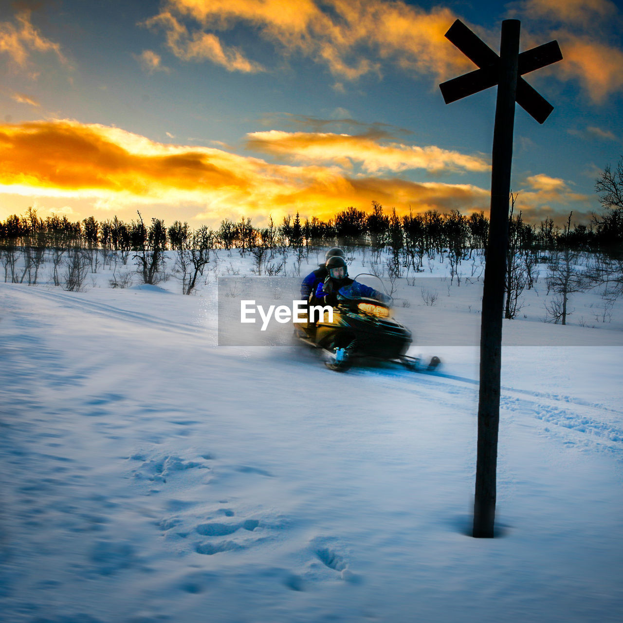 People riding snowmobile on snow covered field against sky during sunset