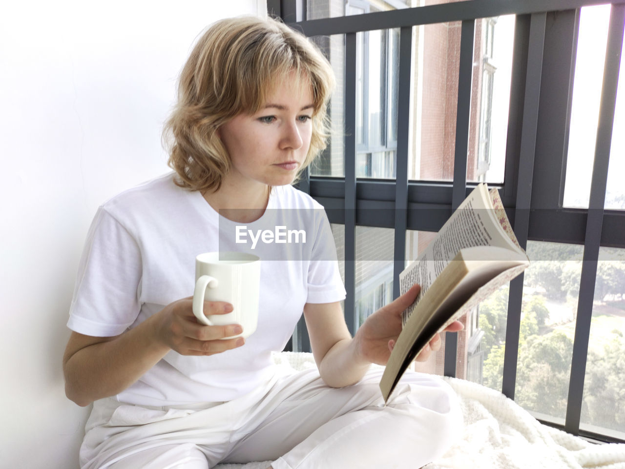 Attractive blond woman sitting on the windowsill reading the book and drinking beverage