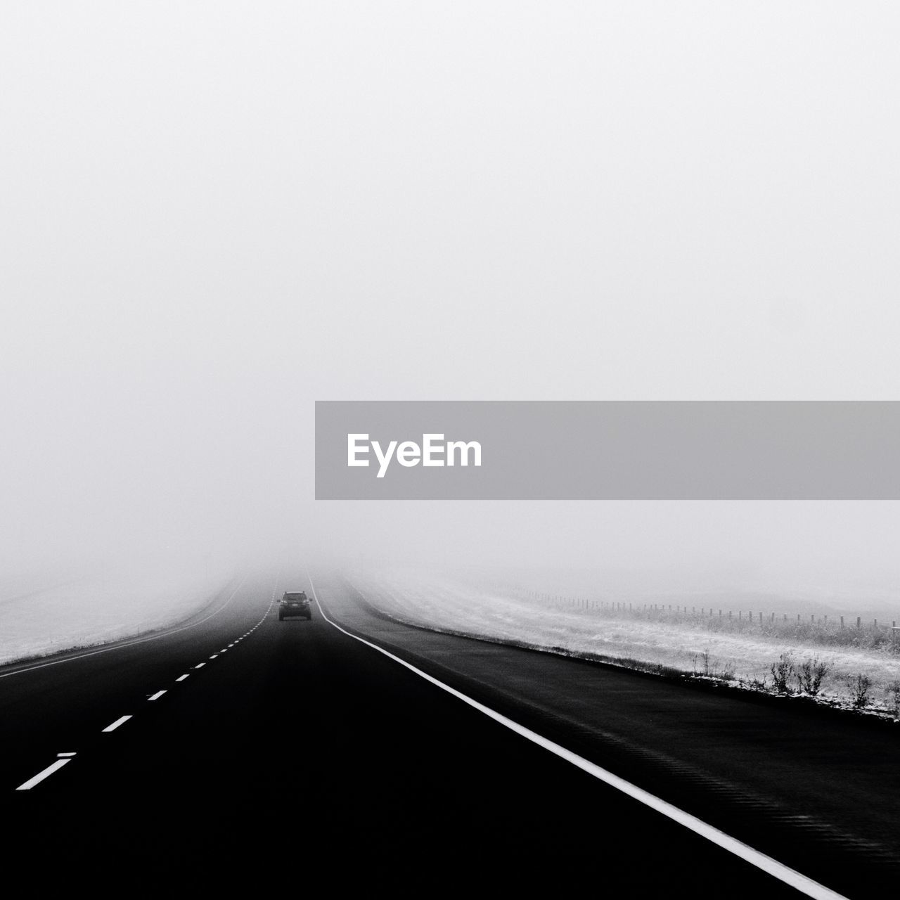 Car moving on road amidst snow covered field against sky in foggy weather