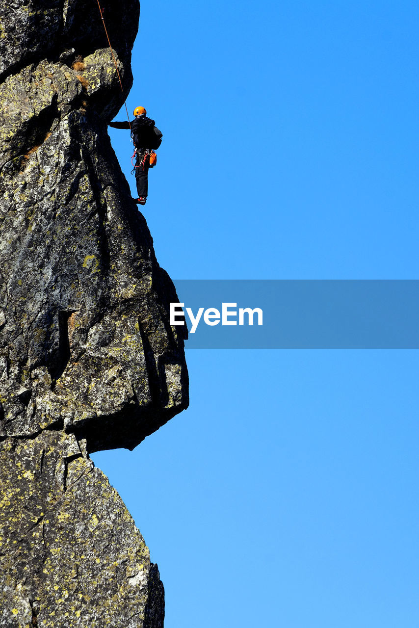 Low angle view of man climbing on rock formation against clear blue sky