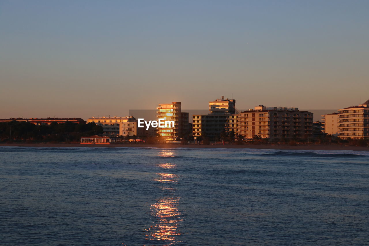 SCENIC VIEW OF SEA BY BUILDINGS AGAINST SKY DURING SUNSET