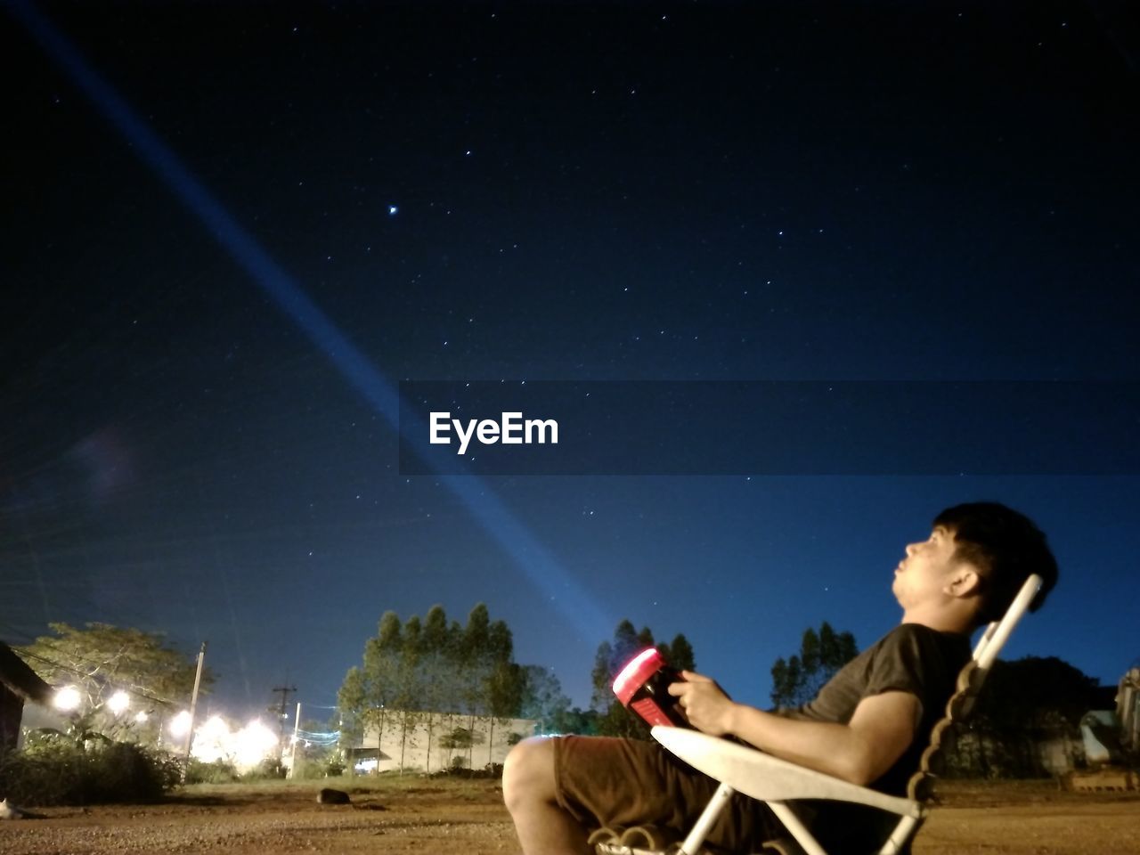 SIDE VIEW OF YOUNG MAN SITTING AGAINST SKY AT NIGHT