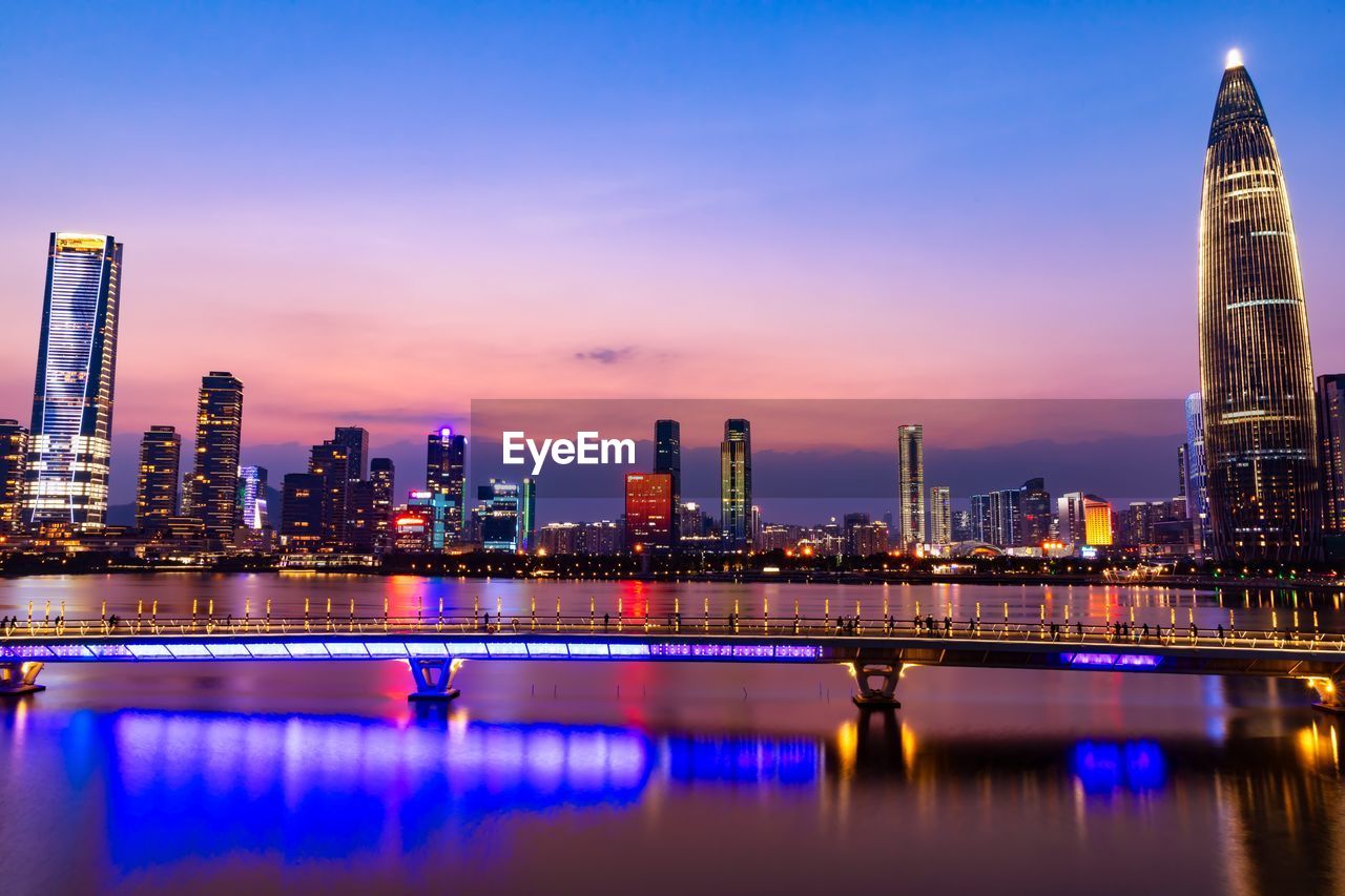 Illuminated buildings by river against sky in city