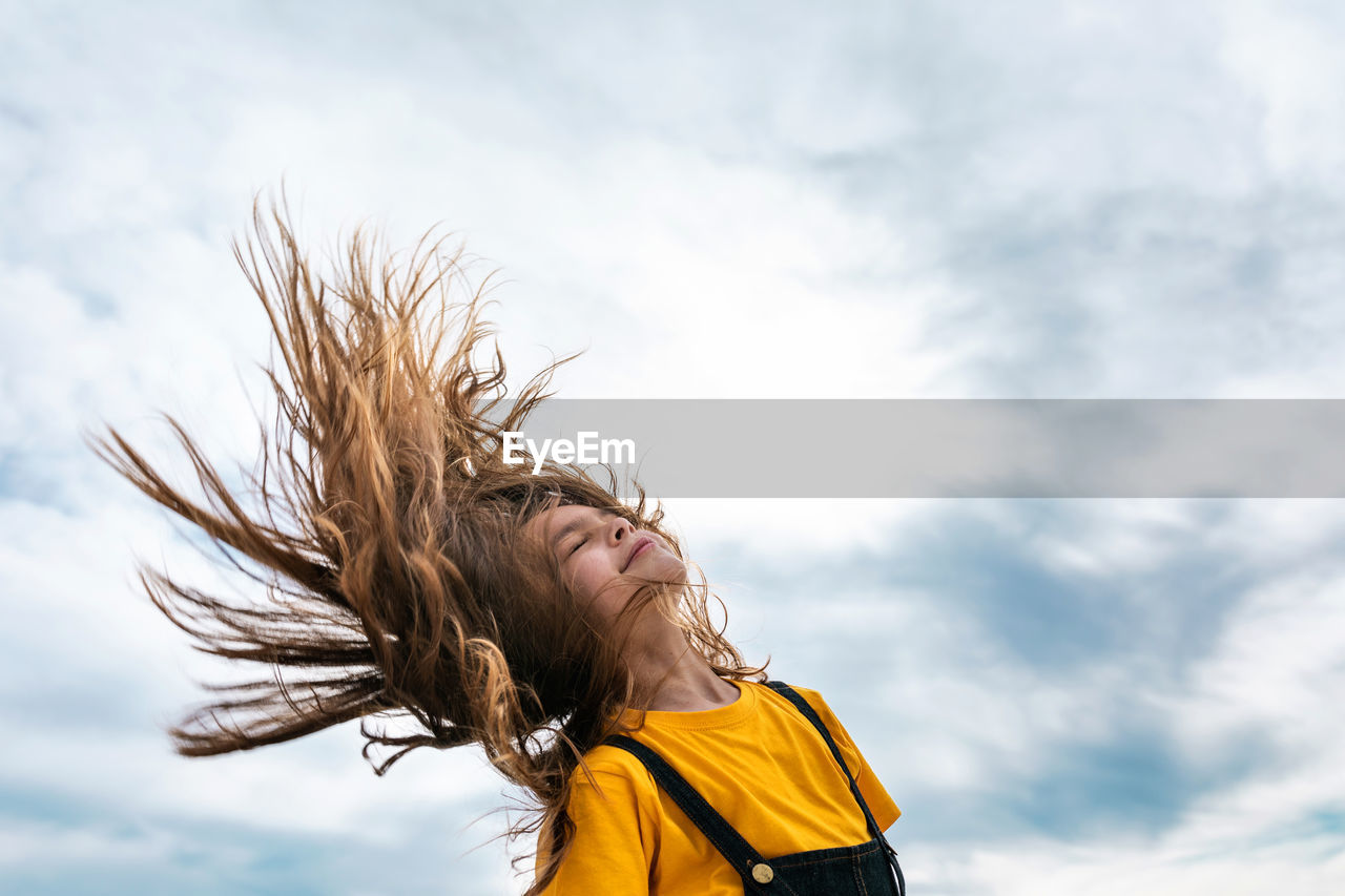 Side view from below of peaceful teenager throwing long hair on background of cloudy sky in summer