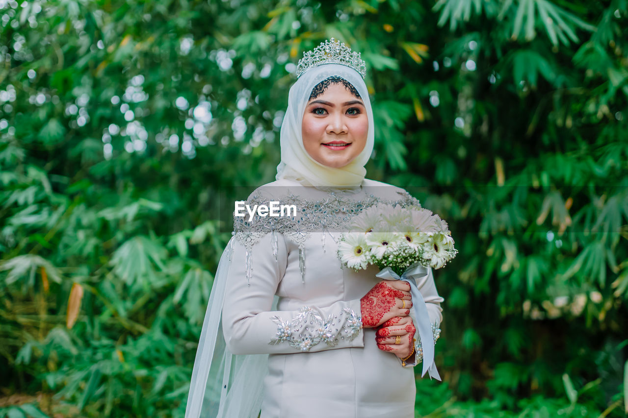 Portrait of smiling bride holding bouquet while standing against trees