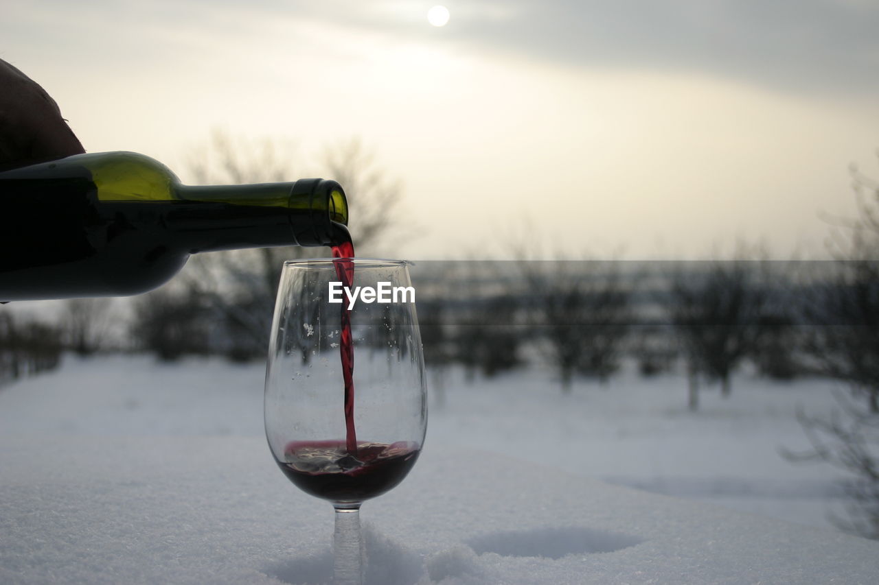 Cropped hand of person pouring red wine in wineglass from bottle during winter at sunset
