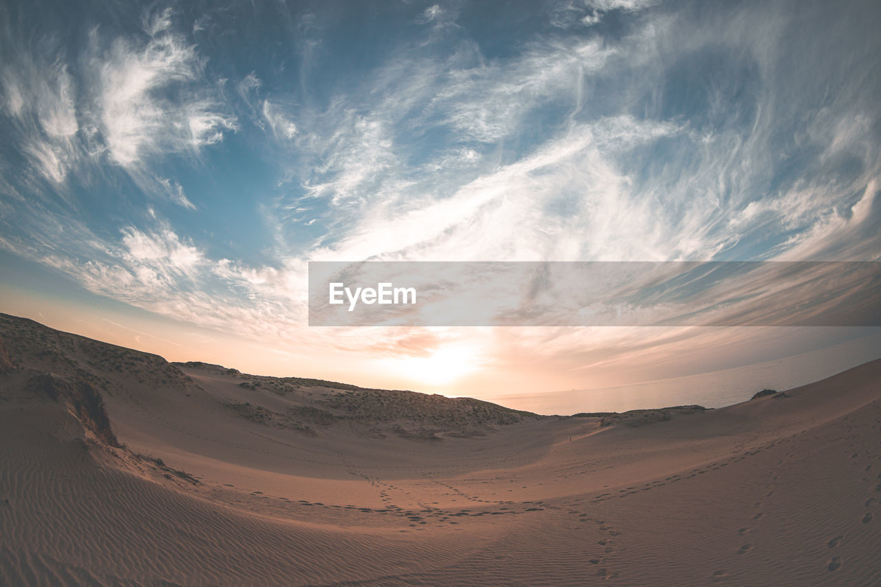 sky, landscape, environment, scenics - nature, sand dune, cloud, land, sand, desert, nature, beauty in nature, natural environment, sunset, sunlight, dramatic sky, climate, tranquility, travel, sun, no people, horizon over land, arid climate, horizon, outdoors, travel destinations, dry, dune, erg, blue, mountain, non-urban scene, tourism, light - natural phenomenon, tranquil scene, singing sand, urban skyline, dusk, summer, drought, panoramic, igniting, orange color, accidents and disasters